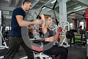 Man trainer helps her slim woman client exercising with heavy dumbbells at the gym