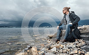 Man traveler with a backpack seets on the seashore against a background of clouds and a mountain range concept of hiking