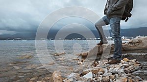 Man traveler with a backpack stands on the seashore against a background of clouds and a mountain range concept of hiking