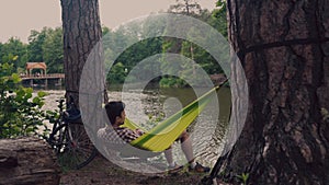 A man traveling by bicycle, resting in a green hammock in the woods by the lake. A cyclist in a hammock at a campsite by