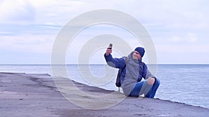 Man traveller makes selfie on smartphone sit on old waterfront at sea in winter.