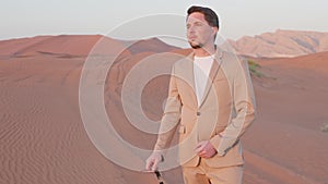 man in trendy suit standing alone in desert, putting off sunglasses and admiring sunset in dunes