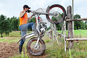 Man with trial mototrcycle training in countryside area, raising bike on rear wheel, leaning against wooden fence
