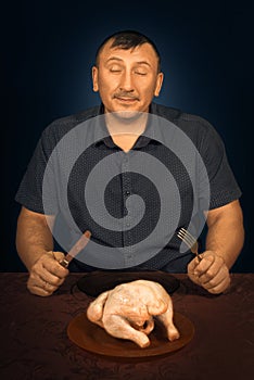 The man tries to eat a crude chicken. Sits at a table with a knife and a forkplug in hands. On a plate food