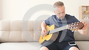 A man tunes an acoustic guitar while sitting on a couch. Resting at home on the sofa