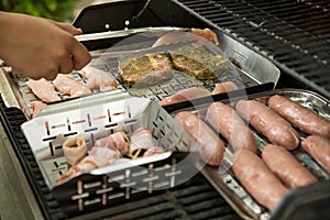 Man turning and roasting chicken filet, pork neck steaks, raw fresh sausages and bacon wrapped dried plums on gas grill BBQ