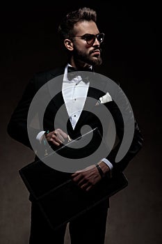 Man in tuxedo holds and opens suitcase while looking away