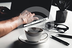 A man types on a laptop, business concept, glasses, a cup of coffee and a pen on a gray background.