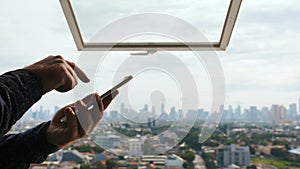 A man types a message on a smartphone against the background of an open window and a panorama of the city of Jakarta