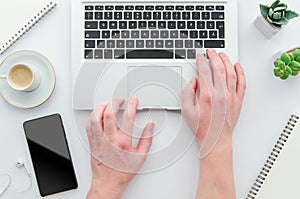 A man is typing on laptop keyboard on modern white office desk with espresso, notepad and blank smartphone screen