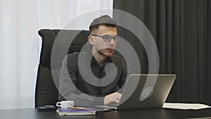 Man typing on laptop keyboard sitting at office. Young confident businessman in glasses works on computer in modern office. Male m