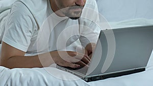 Man typing laptop lying in bed and showing thumbs up, freelancer working online