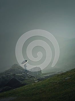 Man with umbrella climbing a mountain hill in a gloomy weather with dense mist. Moody hiking scene with a wanderer walking on the
