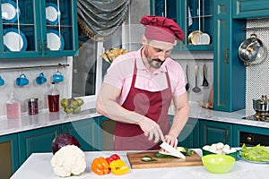 Man in uniform apron cutting zucchini on slice by knife. Orange and red paprika on table prepared for cutting after green zucchini