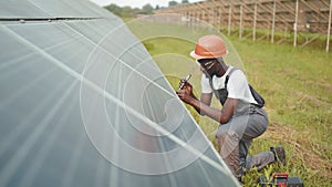 Man in uniform and helmet using screwdriver for repairing solar panels on station. Installation of photovoltaic cells