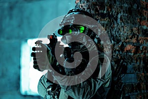 Man in uniform with machine gun and turned on night vision device beside brick wall. Closeup airsoft soldier with green light on