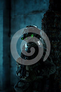 Man in uniform with machine gun and turned on night vision device move between walls Airsoft soldier with green light on face in photo