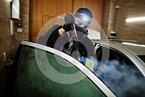 Man in uniform and respirator, worker of car wash center, cleaning car interior with hot steam cleaner. Car detailing concept