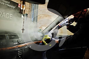 Man in uniform and respirator, worker of car wash center, cleaning car interior with hot steam cleaner. Car detailing concept