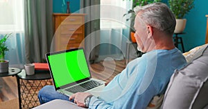 Man Uses Laptop with Green Mock up Screen Sitting on a Couch at Home. An