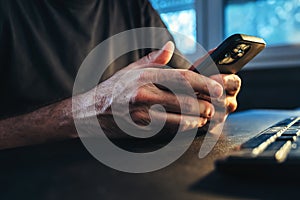 Man using cell phone at home office in low key interior. Casual male typing text message on smartphone while sitting at the desk