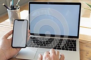 Man using computer holding smartphone with white empty mockup screen