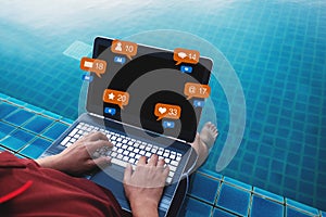 A man using computer laptop at poolside in summer holiday, with social media, social network notification icons