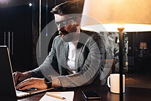 Man using contemporary mobile notebook. Bearded businessman working at night in modern loft office. Project manager sitting at woo photo