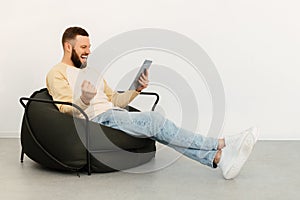 Man Using Digital Tablet Shaking Fists Sitting In Chair Indoors