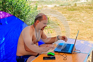 Man using his laptop sitting at a folding picnic table in a camping. Working while traveling. Freelance work