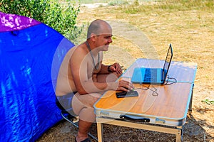 Man using his laptop sitting at a folding picnic table in a camping. Working while traveling. Freelance work.