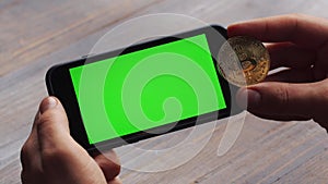 Man using horizontal smartphone with green screen. Close-up shot of man`s hands with mobile phone and a gold bitcoin