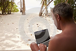Man using a laptop computer on a beach, over shoulder view