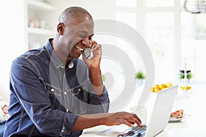 Muž prenosný počítač a hovorenie na telefón v kuchyňa na 
