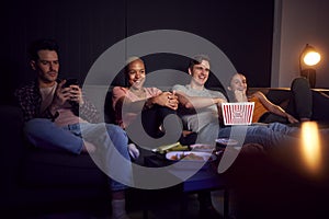 Man Using Mobile Phone Whilst Friends Watch TV At Home In Evening