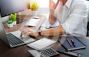 A man using mobile smartphone communicates on internet technology with typing keyboard of laptop computer on office desk.