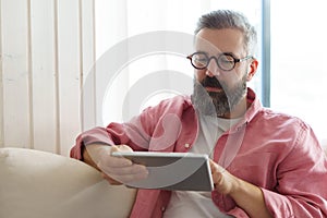Man using smart thermostat, adjusting, lowering heating temperature at home. Man watching tv show on tablet, video