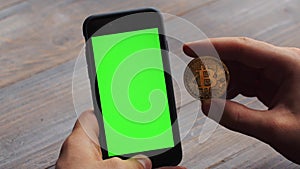 Man using vertical smartphone with green screen. Close-up shot of man`s hands with mobile phone and a gold bitcoin coin