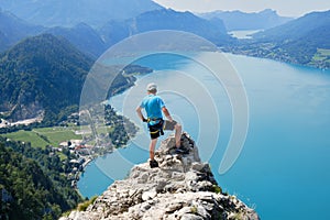 Man with via ferrata gear climbs on top of a rock above Attersee, Austria. Climber, lake, turquoise, summer, tourism, adventure.