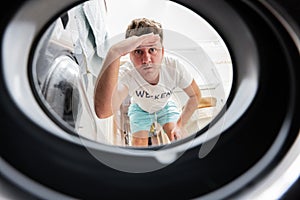 Man view from washing machine inside. Male does laundry daily routine. Hand over forehead looking far away, watching or searching