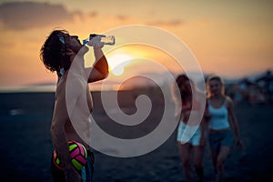 Man with volleyball at the beach drinking water, at sunset. Preparing to play volleyball with friends. Sport, recreation, fun,