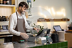 Man waiter working in coffee shop using terminal while standing at counter