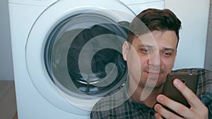 Man is waiting the washing machine with bedspread and talking on smartphone.