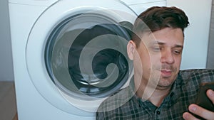 Man is waiting the washing machine with bedspread and talking on smartphone.