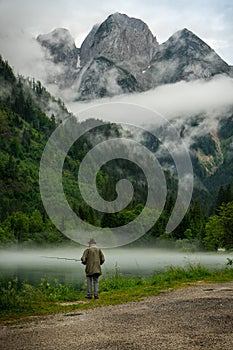 Man walking alone on a riverbank, with a rod in one hand and a net in the other, on a foggy morning