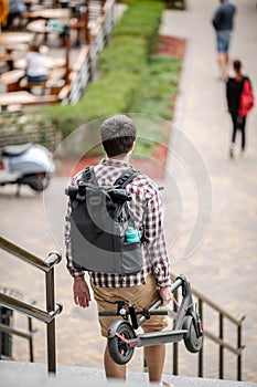 Man walking with folded electric scooter up stairs in city near modern building. Ecological technological lifestyle. E-Mobility.