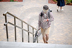 Man walking with folded electric scooter up stairs in city near modern building. Ecological technological lifestyle. E-Mobility.