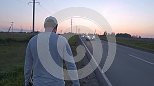 A Man Walking On The Side Of A Car Road, View From The Back. Cars Passing Nearby, Beautiful Shot at Sunset. The Concept