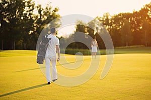 A man walks along the golf course with a golf club bag and heads towards the girl who is standing in front of him