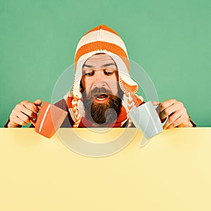 Man in warm hat holds cyan and orange cup on green and yellow background. Hipster with beard and curious face having